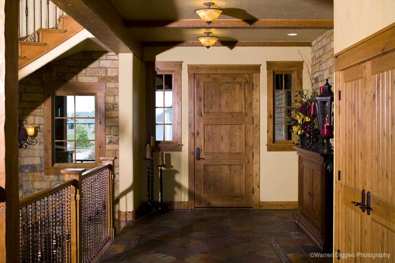 Rustic entryway with stone wall, wood-paneled door, windows, and ornate staircase railing.