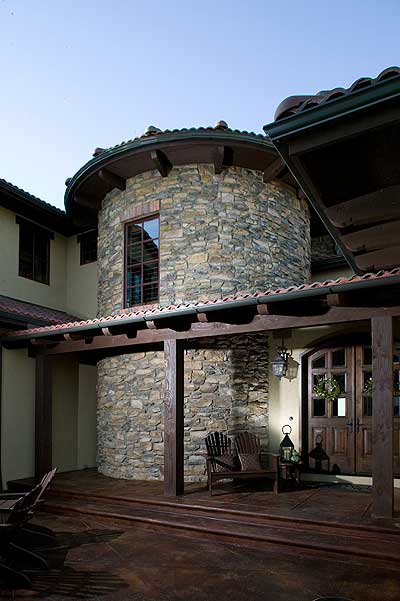 House plan exterior featuring a round stone tower with a single window, covered porch with wood beams, and double wooden doors.