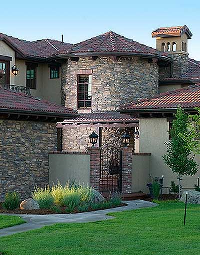 House plan exterior showing a stucco and stone building with a rounded tower and tiled roof. Features a decorative iron gate.