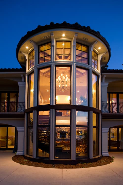 House plan exterior featuring a multi-story rounded entryway with extensive glass, wrought iron spiral staircase, and chandelier.