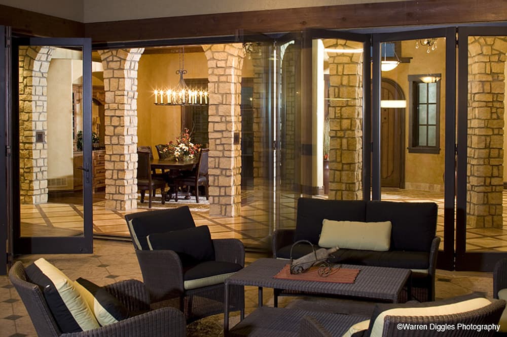 Exterior view of house plan with stone columns, arched doorways, and large folding glass doors opening to a patio.