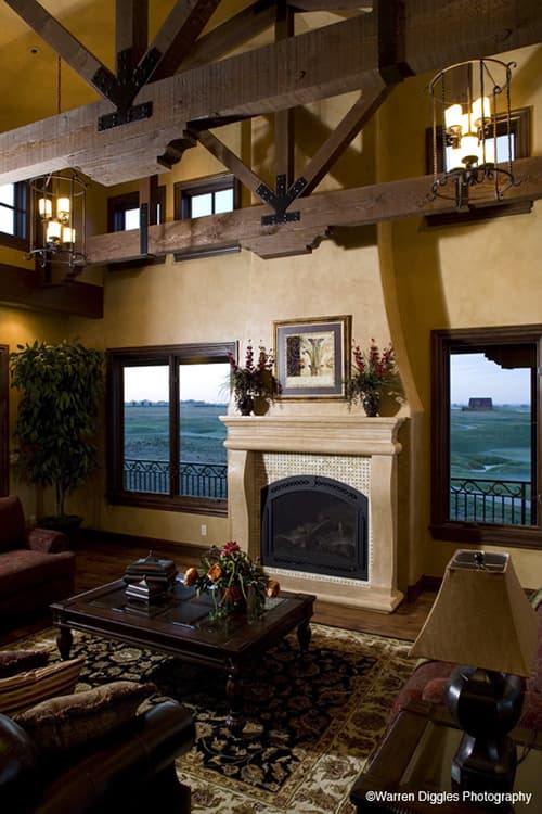 Interior view of living room with exposed wooden beams, stone fireplace, and large windows overlooking a landscape.