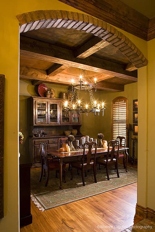 Dining room with large wooden table, hutch, chandelier, beamed ceiling, and arched brick entryway.