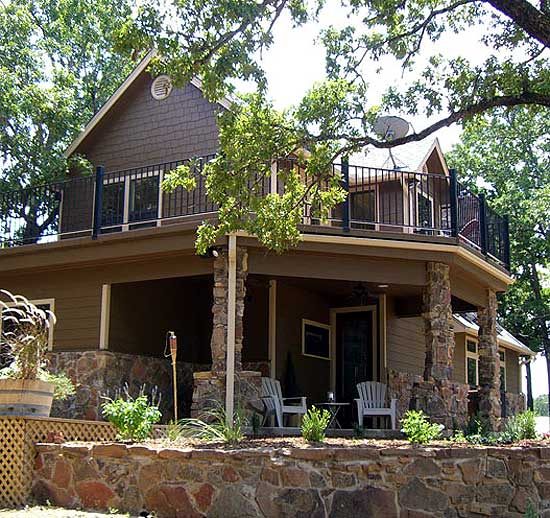 House plan exterior: Two-story home with shingle siding, upper balcony, stone-clad porch supports, and wide gable roof.