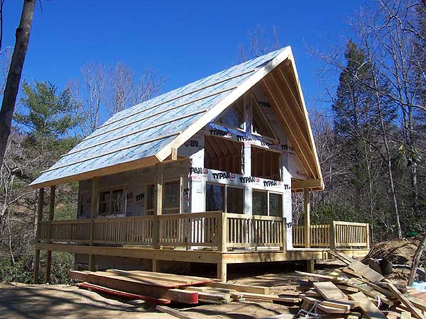 House plan exterior of a one-story cabin with a wraparound porch, gabled roof, and exposed rafters during construction.